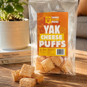 Bag of Three Yellow Dogs Yak Cheese Puffs standing on a wood table with several puffs piled in front of it.